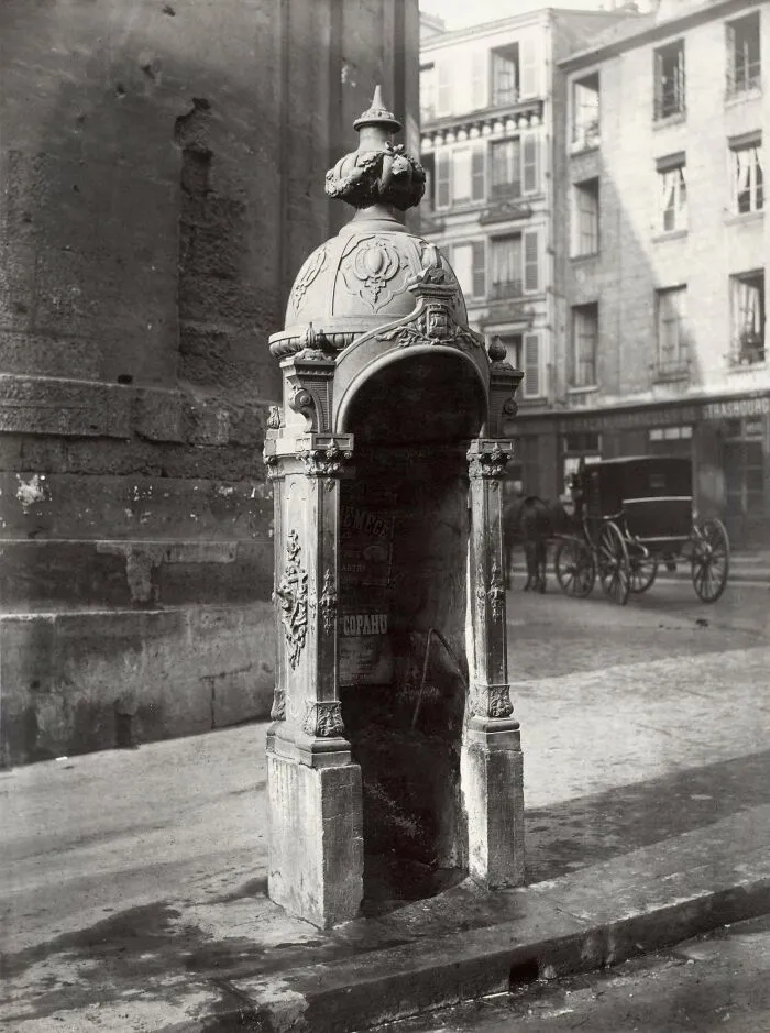Ornate historic site water fountain in a city street with vintage architecture and horse-drawn carriage in background.