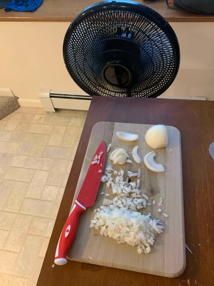 Chopped onion on a wooden cutting board with a red knife nearby and a fan blowing on the cutting area.