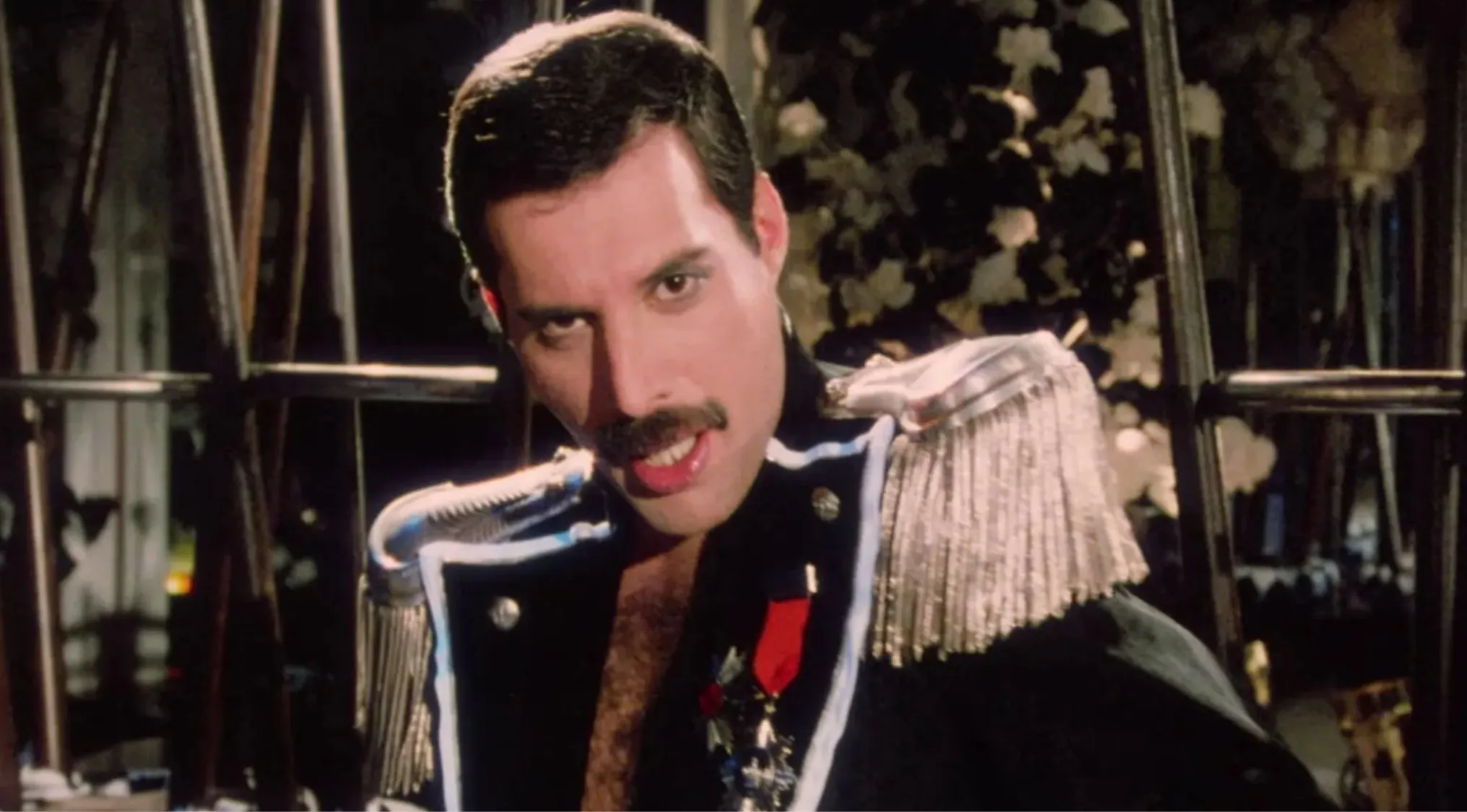 Freddie Mercury performing on stage in a decorated military-style jacket, lead singer of iconic rock band Queen.