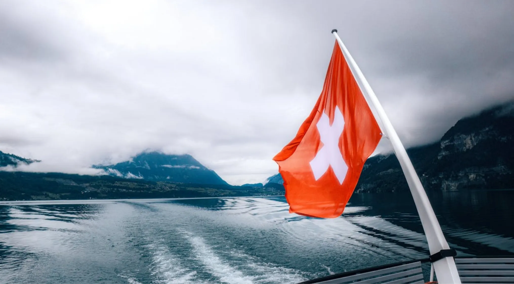 Swiss flag waving on a boat with mountains and lake in the background representing currency in Switzerland trivia night.
