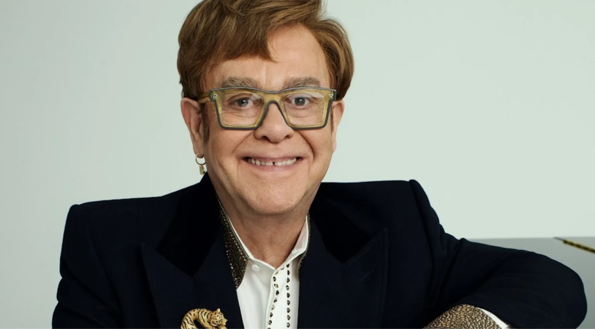 Elton John smiling, wearing glasses and a black jacket with a decorative pin, posing against a plain light background