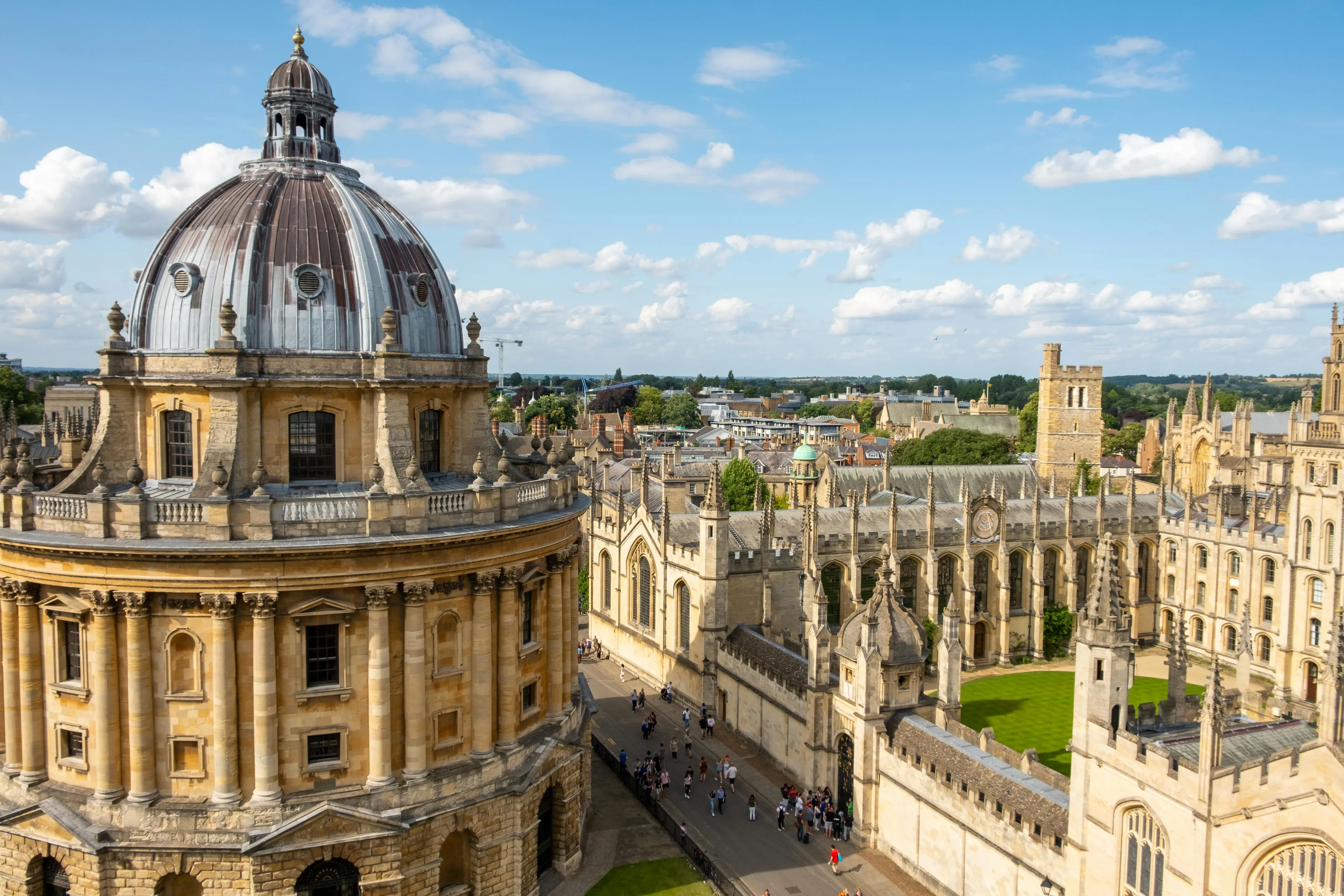 Radcliffe Camera and historic buildings at Oxford University on a sunny day with scattered clouds and visitors walking.