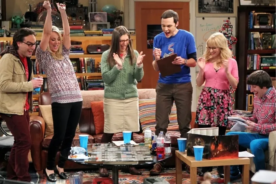 Cast of The Big Bang Theory playing a board game in an apartment, celebrating a winning move together.