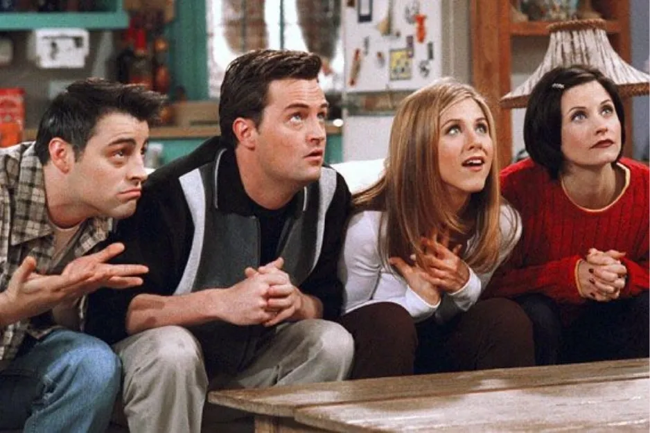 Friends cast sitting closely on a couch in a living room, engaged and looking intently at something off-camera.