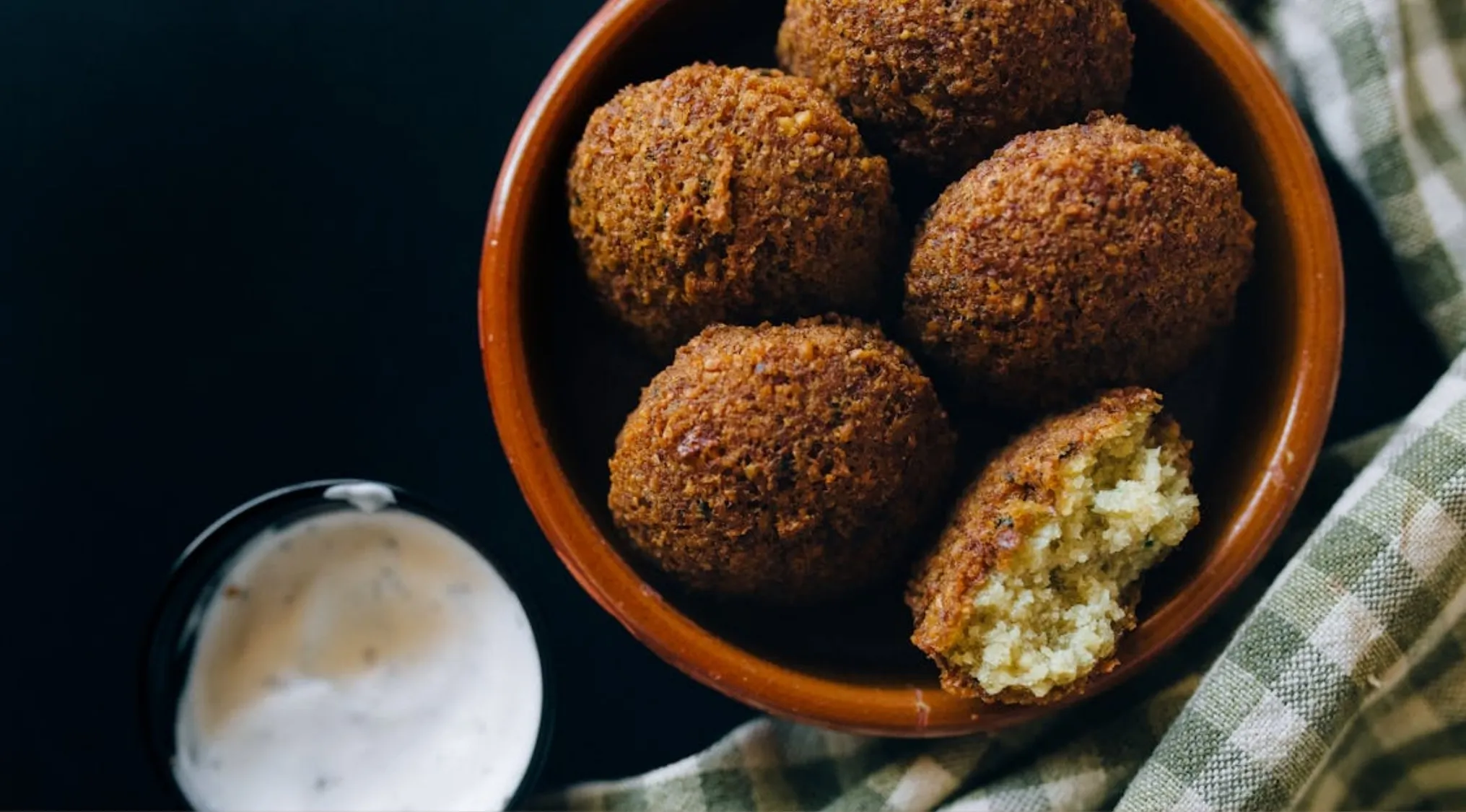 Bowl of crispy falafel balls with creamy dipping sauce and a green checked napkin on a dark background