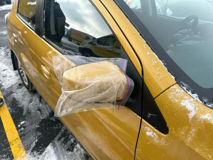 Yellow car with a broken side mirror temporarily fixed using a plastic bag filled with insulation foam in a snowy parking lot.