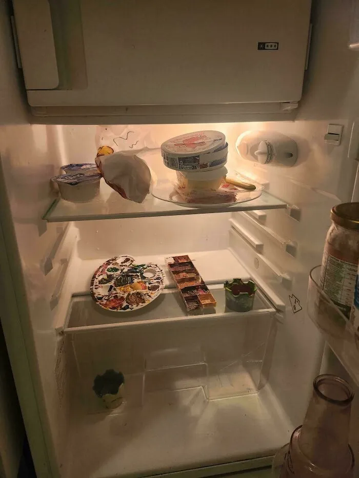 Partially empty fridge interior with art supplies including a paint palette, watercolor set, and a few food items inside.