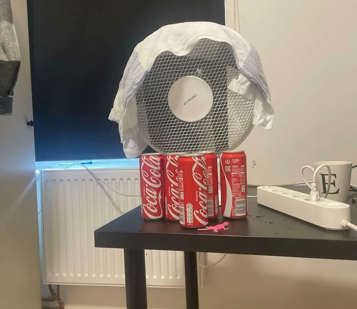 Fan propped up by Coca-Cola cans with a towel draped over it, a creative example of if it's stupid but it works.
