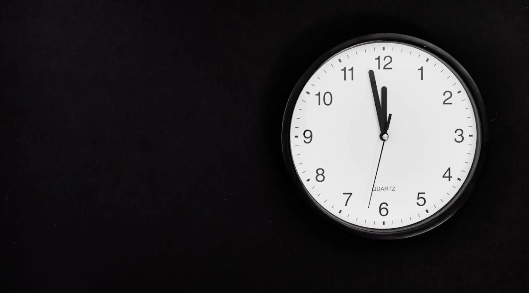 Round white wall clock showing time at 12:57 with black frame on a black background for testing basic math skills.