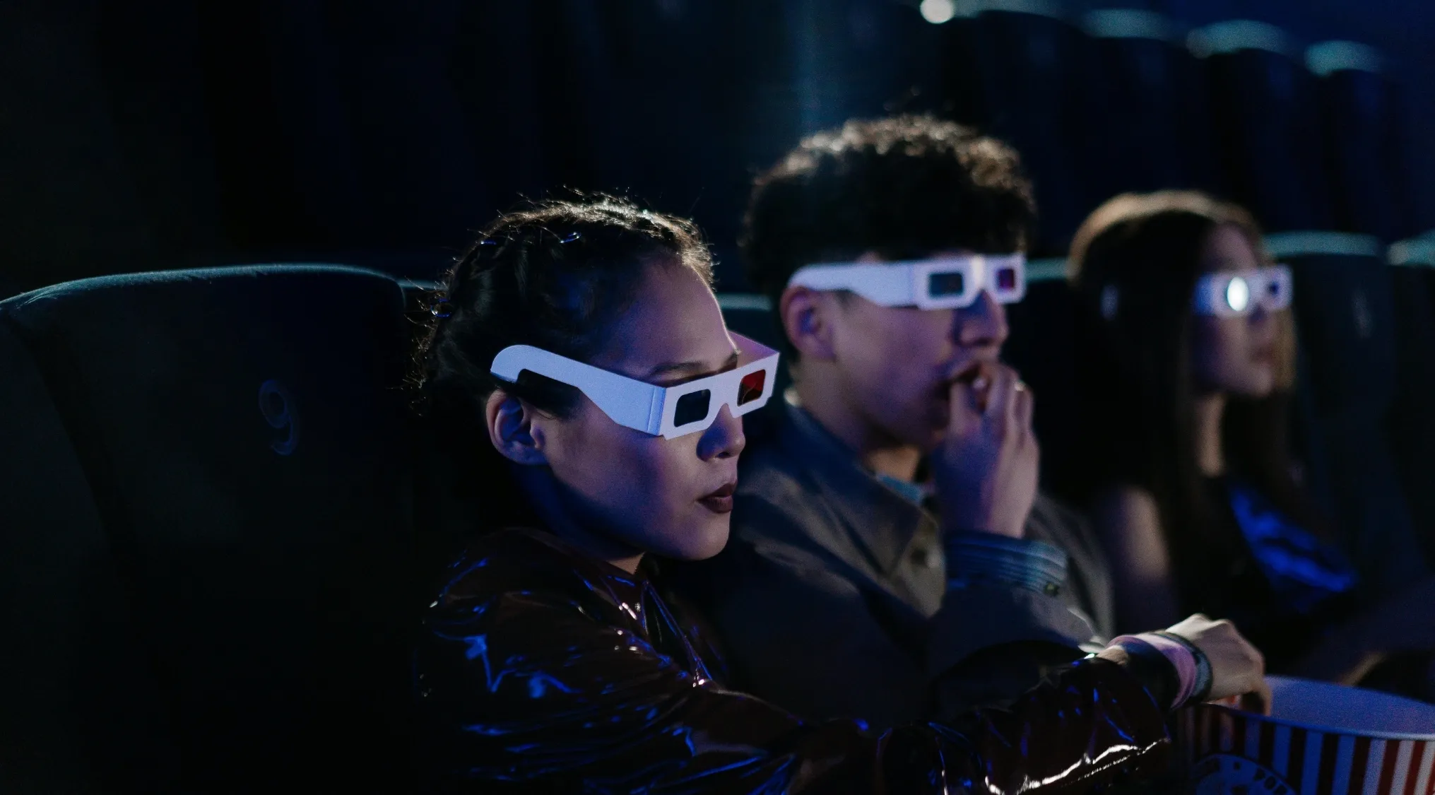 Three people wearing 3D glasses watching a movie in a dark theater, enjoying a cinephile movie quiz anniversary edition.