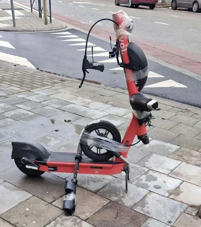 Damaged electric scooter on a sidewalk, wrapped with tape, one wheel detached, weird and unhinged things seen on Brussels streets.