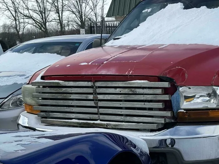 Red truck with a makeshift grill repair made of metal bars covered partially in snow in a parking lot.