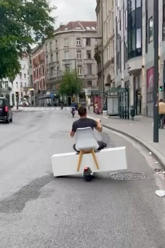 Man riding a homemade motorized vehicle with a chair and a white box on the streets of Brussels, showing weird street scenes.