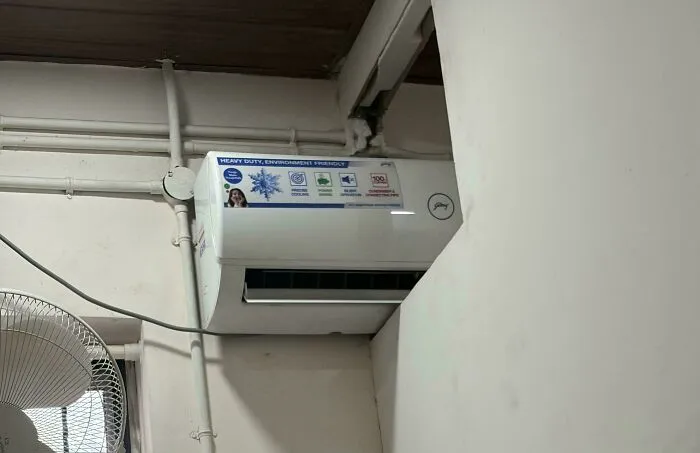 Air conditioner mounted high on a wall with visible pipes and a circular fan below in an indoor setting, example of if it works.