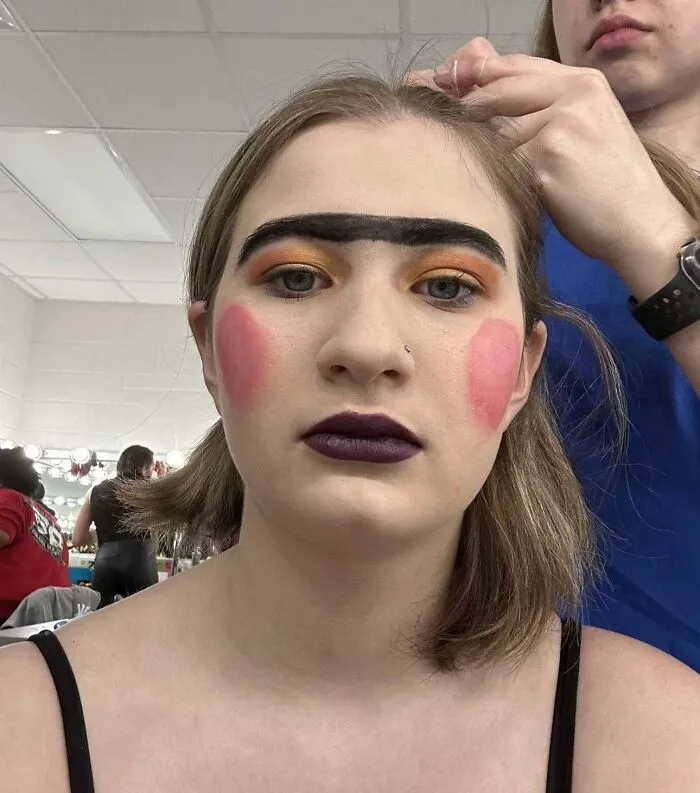 Young woman with stage makeup featuring bold unibrow, bright blush, dark lipstick, and orange eyeshadow during backstage preparation.