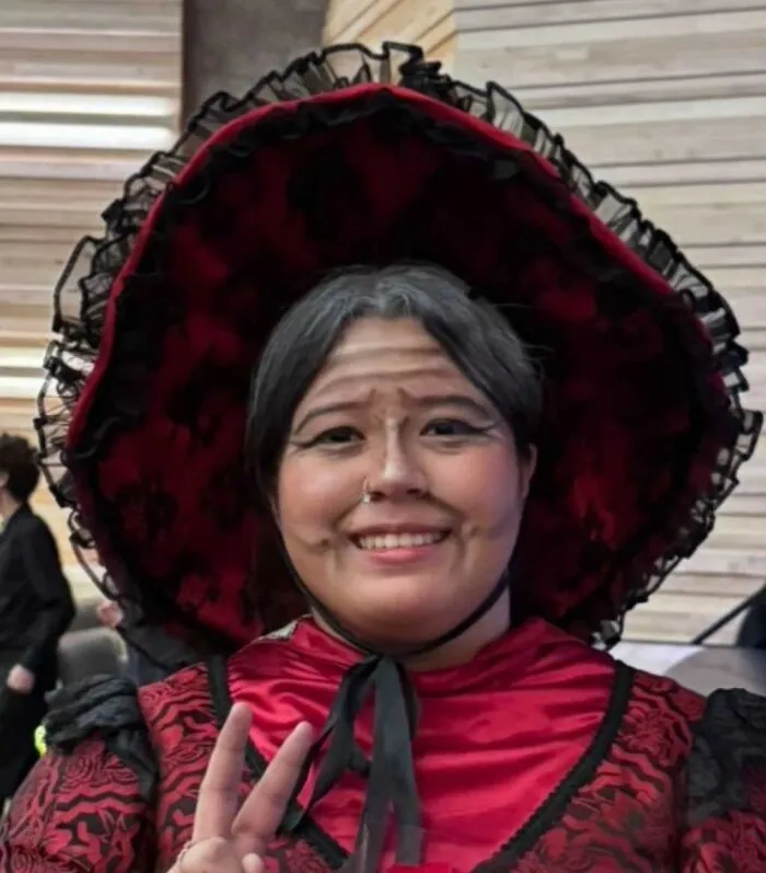 Person wearing stage makeup with exaggerated wrinkles and a large red and black costume hat posing with a peace sign.