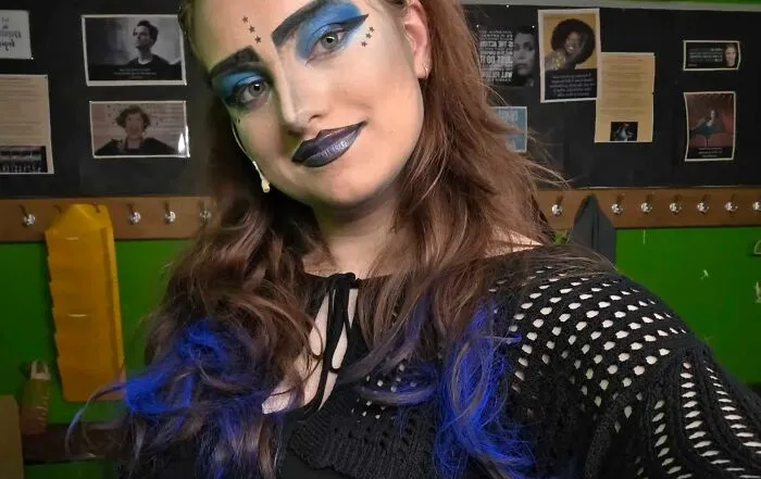 Young woman with magical stage makeup featuring bold blue eyeshadow and dark lips, posing in a backstage dressing room.