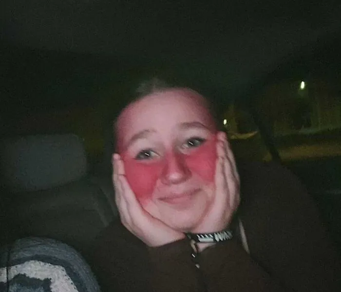 Person with exaggerated stage makeup in red tone posing with hands on cheeks in dimly lit environment showing stage makeup effects.