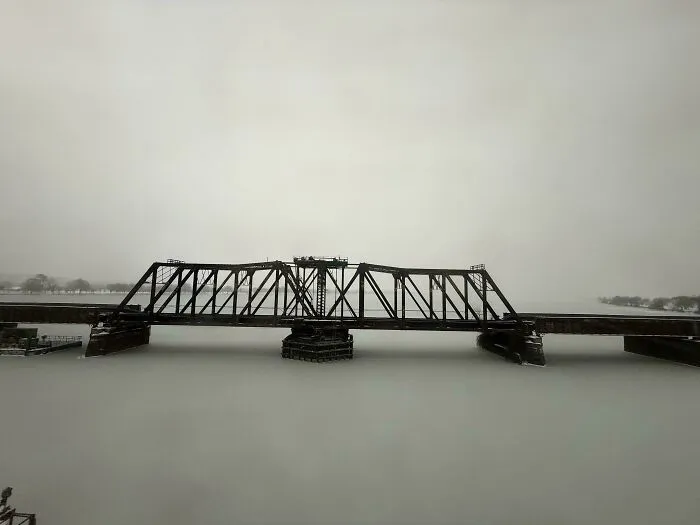 Steel railroad bridge covered in snow with a snowstorm hitting the U.S., creating a winter landscape and icy river below.