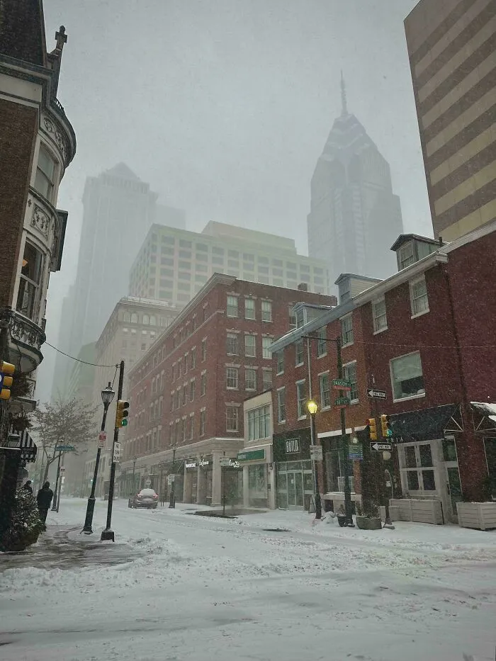 Snowstorm hitting U.S. city streets with heavy snow, reduced visibility, and people walking in winter weather conditions.