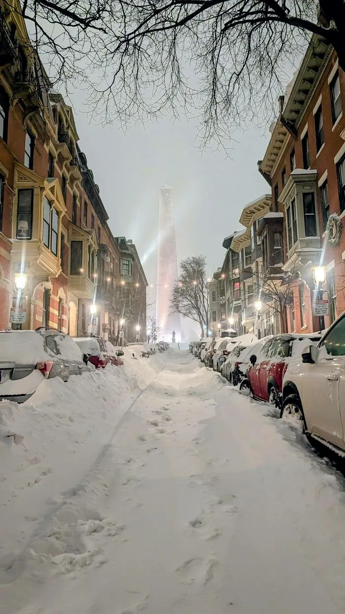 Snowstorm hitting the U.S. with heavy snow covering streets, cars, and buildings around a tall monument at night.