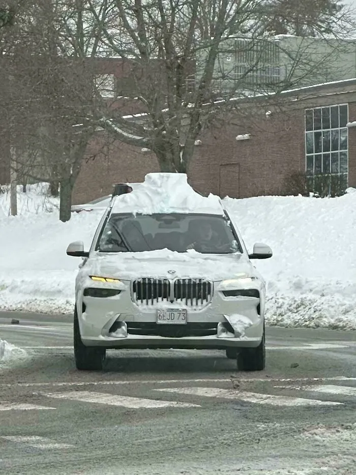White BMW SUV covered with snow on a city street during a heavy snowstorm hitting the U.S. in winter weather conditions.