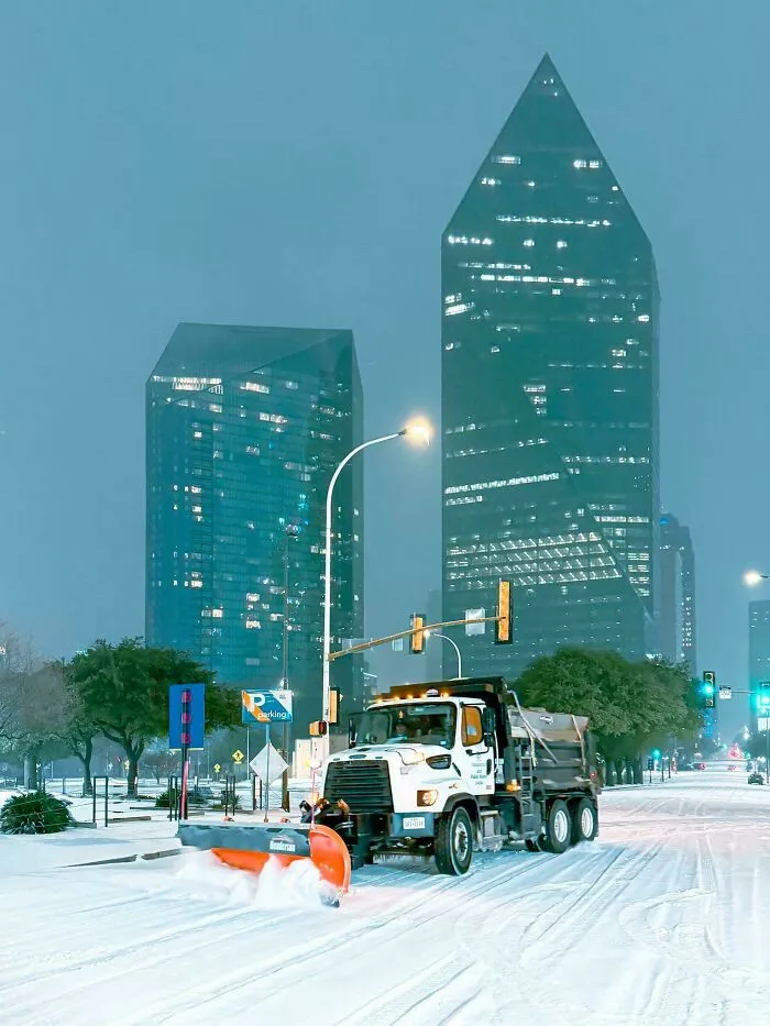 Snowplow clearing snow on city streets during a heavy snowstorm hitting the U.S. with tall buildings in the background.