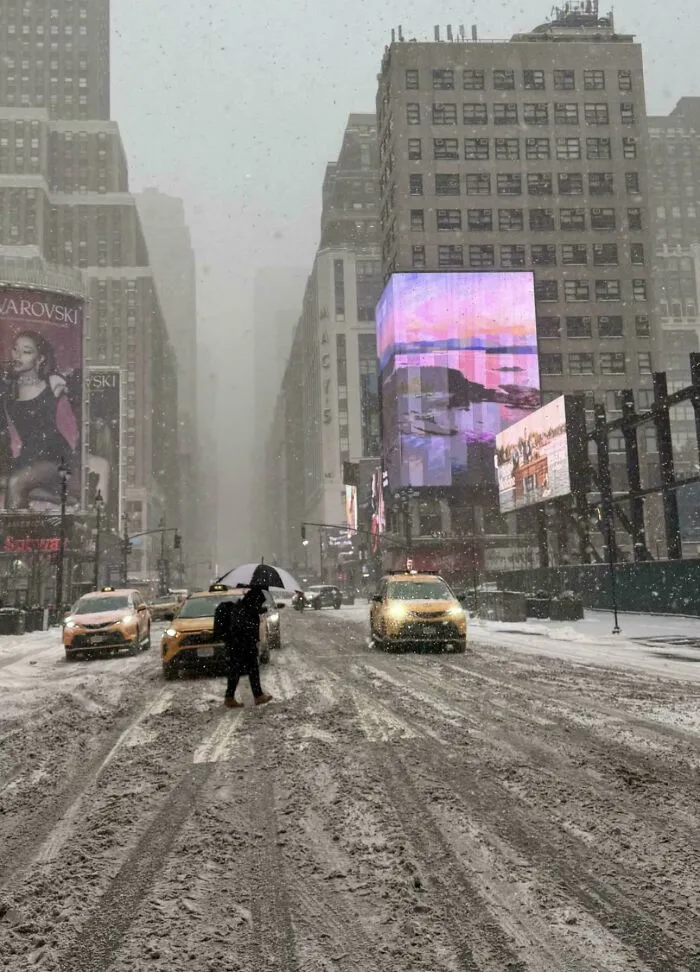 Snowstorm hitting the U.S. city with heavy snowfall, taxis, and a pedestrian crossing the snowy street under an umbrella.