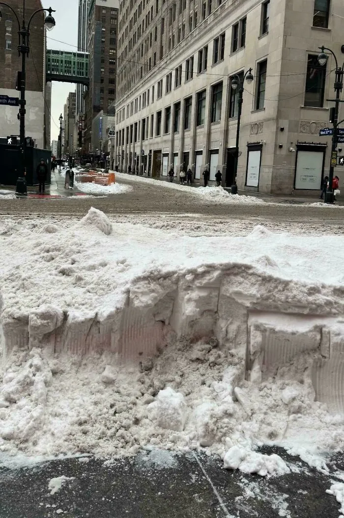 Snowstorm hitting U.S. city street with large snow piles and slushy roads amid tall urban buildings on a winter day.