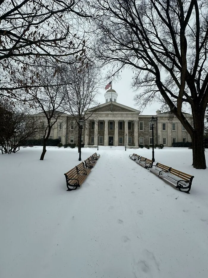 Historic building framed by barren trees with snow-covered benches and ground after a heavy snowstorm in the U.S.
