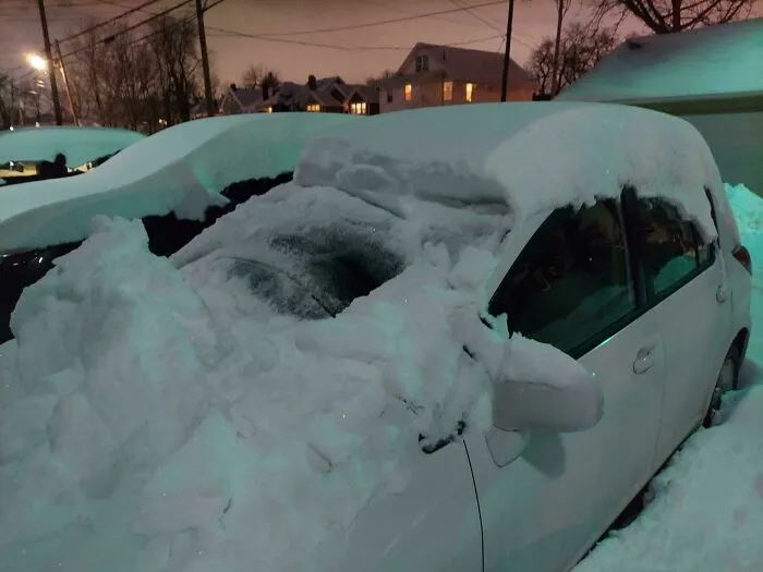 Car covered in heavy snow during a snowstorm in the U.S. at night, with houses and trees in the background.