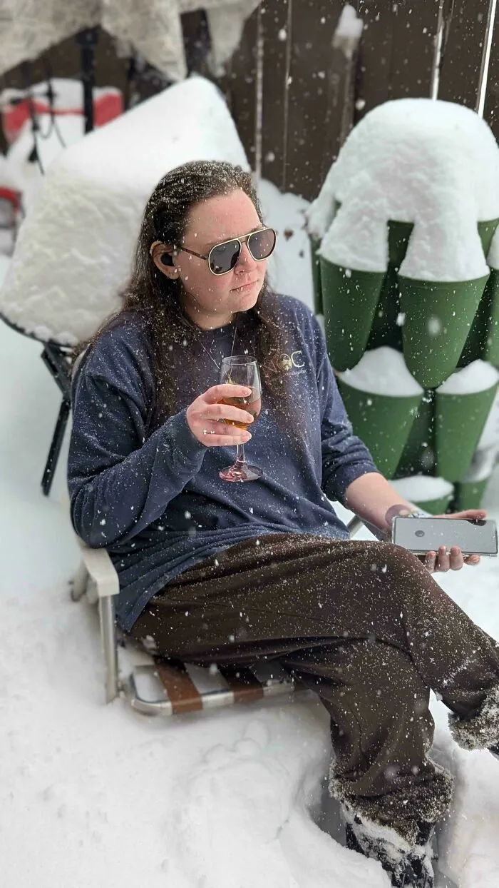 Person wearing sunglasses sitting in snowstorm outdoors, holding a drink and phone amidst heavy snow accumulation.