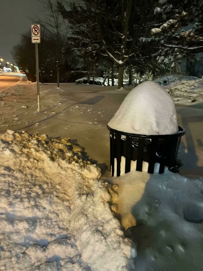 Snowstorm hitting the U.S. with heavy snow covering a trash can and the surrounding sidewalk at night.