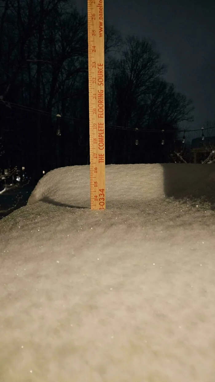 Snowstorm hitting the U.S. with snowfall over 18 inches measured by a wooden ruler at night near bare trees.