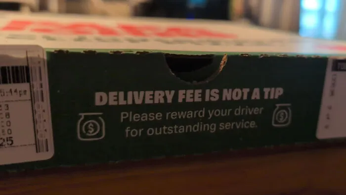 Close-up of pizza box warning that delivery fee is not a tip, highlighting issues related to consumerism and service industry.