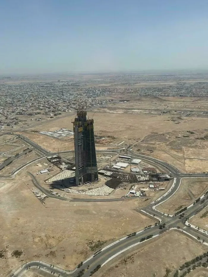 A dystopian urban hell scene showing a lone skyscraper under construction surrounded by barren land and sparse buildings.