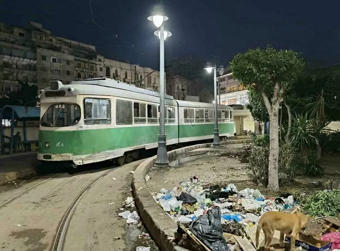 Old green tram at night near trash pile and urban buildings, depicting dystopian urban hell and modern life challenges.
