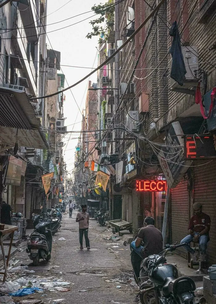 Narrow urban street filled with tangled wires, motorcycles, litter, and people, showing a dystopian urban hell environment.