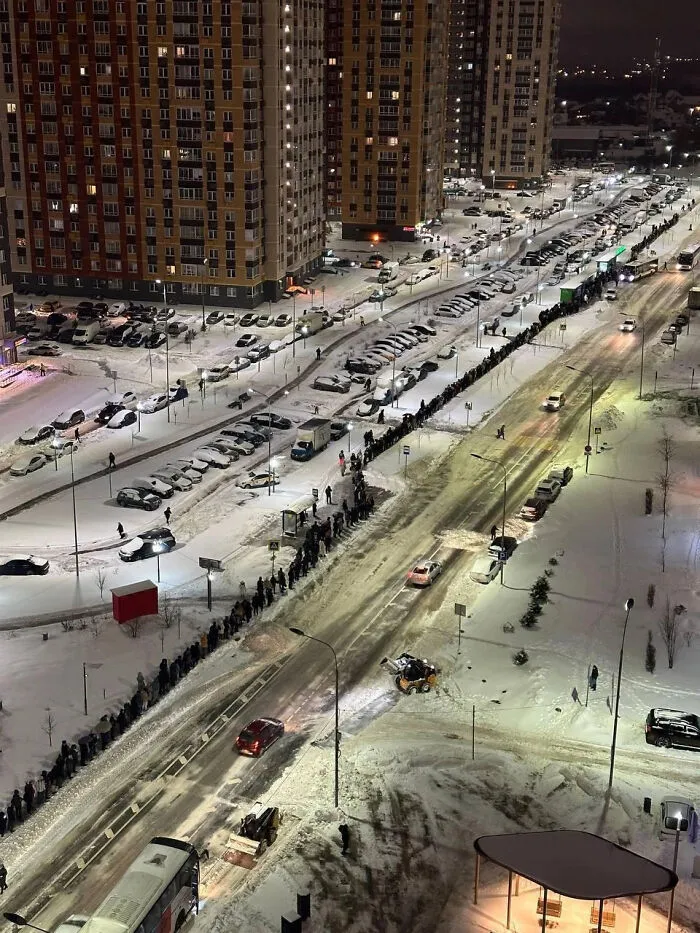 Long line of people along snowy urban street beside high-rise apartments at night, showing dystopian urban hell atmosphere.