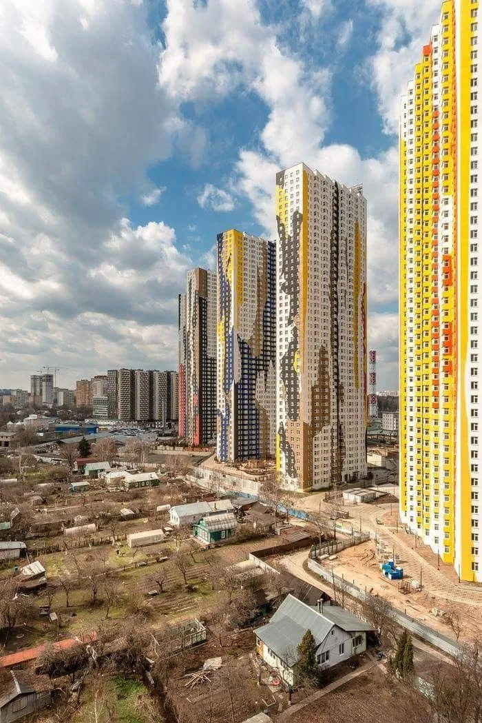 High-rise apartment buildings tower over small rural houses in a dystopian urban hell cityscape under a cloudy sky.