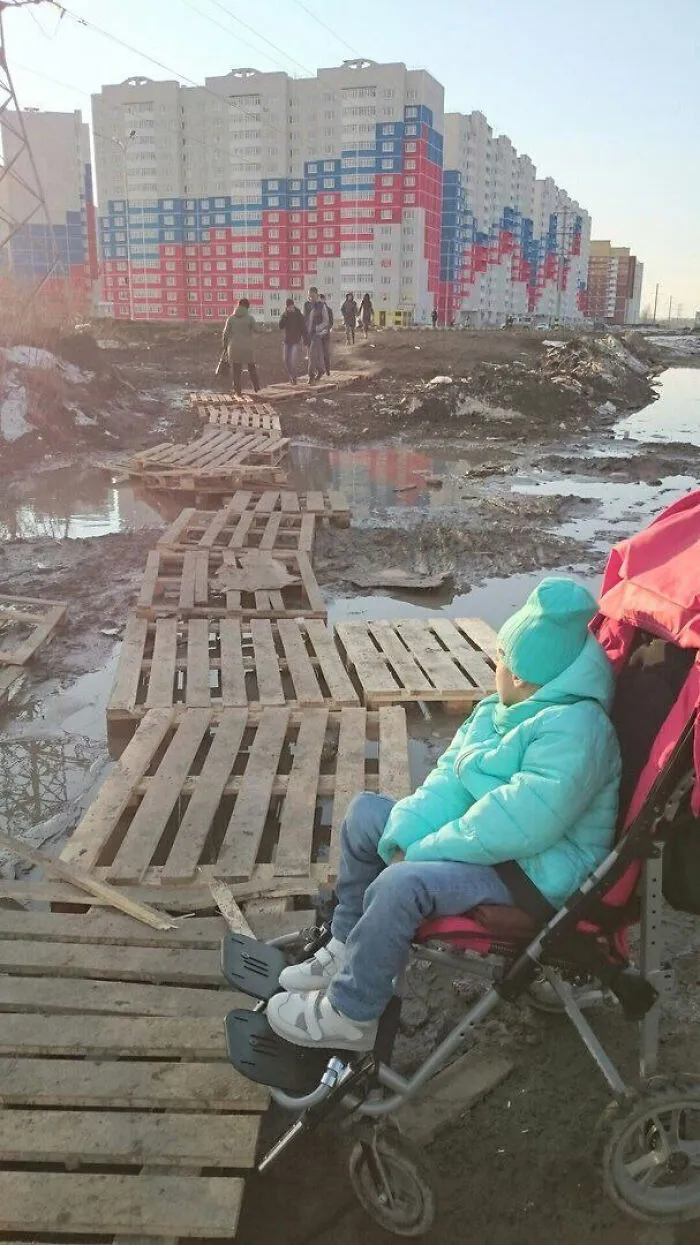 Child in a stroller looks at makeshift wooden path over muddy urban hell near deteriorating apartment buildings.