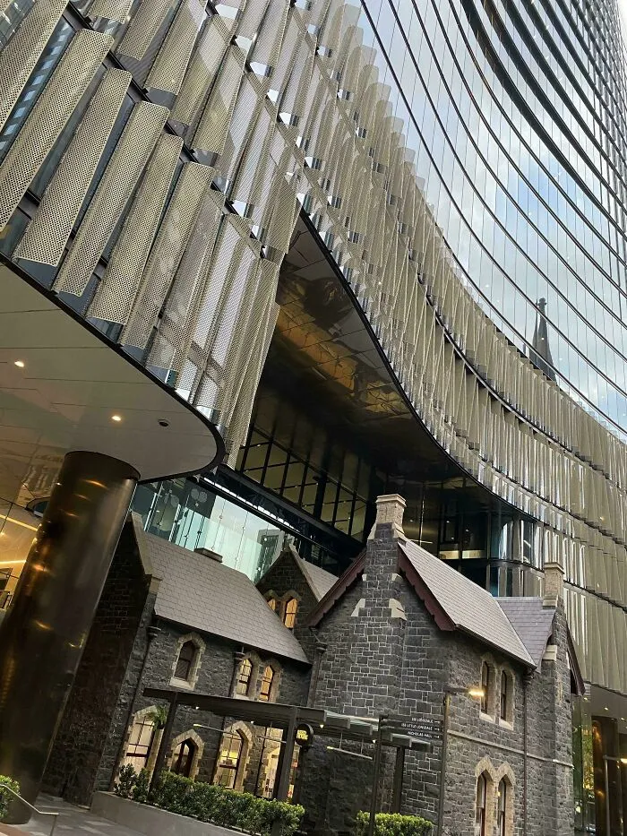 Old stone building trapped beneath modern glass skyscraper in a dystopian urban hell scene showing the price of modern life.