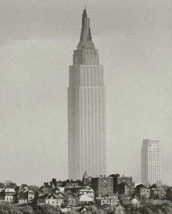 A sepia-toned image of an extremely large skyscraper towering over smaller buildings, creating a scary and eerie scale.