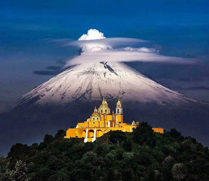 Beautiful architecture of a vibrant yellow church nestled on a hill with a majestic, snow-capped mountain in the background.