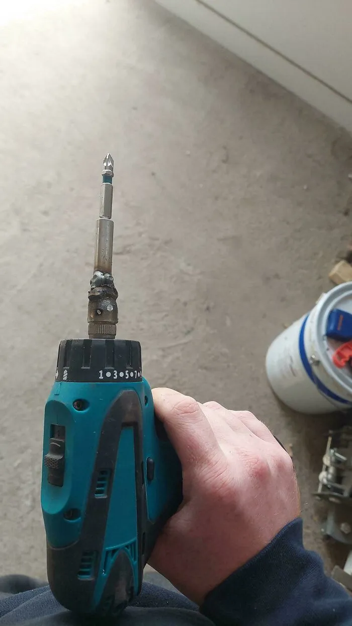 Hand holding a power drill with a homemade welded extension and screwdriver bit in a redneck engineering style.