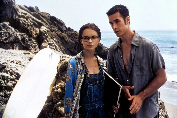 Two young adults on a rocky beach, reflecting on ’90s childhood memories with surfboard and casual clothing.