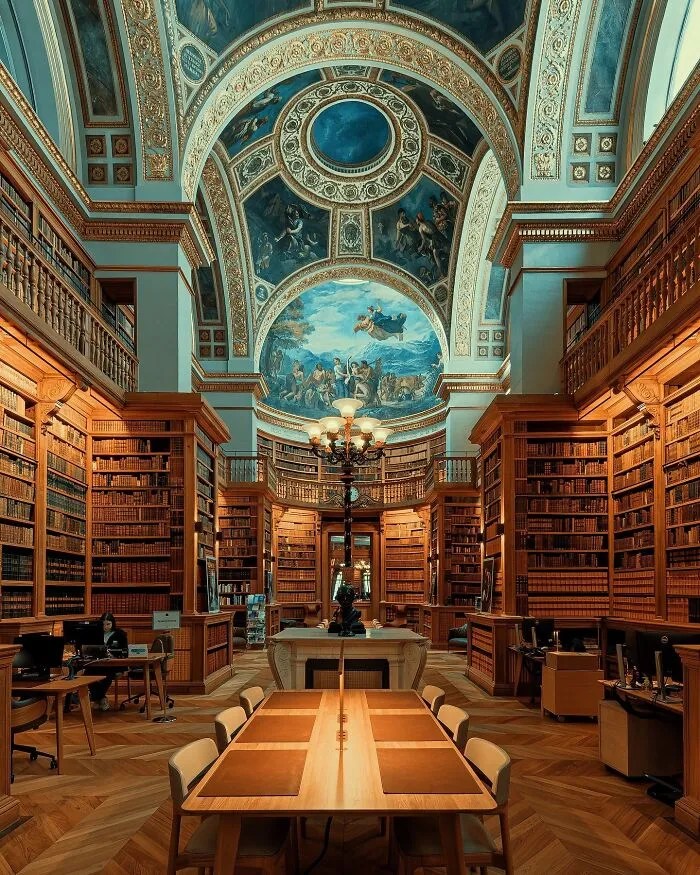 Grand library with towering bookshelves, intricate ceiling art, and a long central table showcasing beautiful architecture.