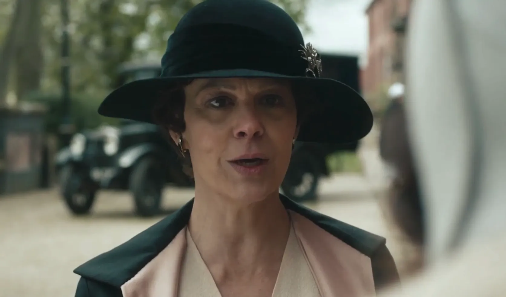 Close-up of a woman wearing a black hat, speaking outdoors in a historical setting, related to receptionist office administrator role.