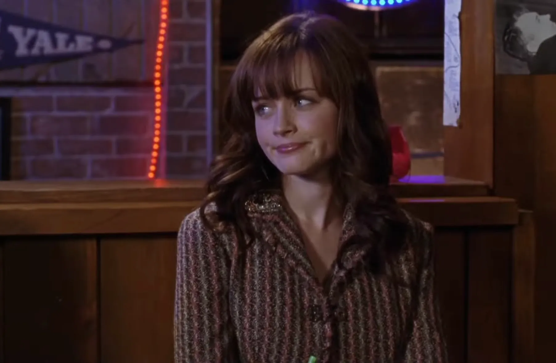 Rory Gilmore sitting indoors with a thoughtful expression, wearing a patterned jacket with wavy brown hair.