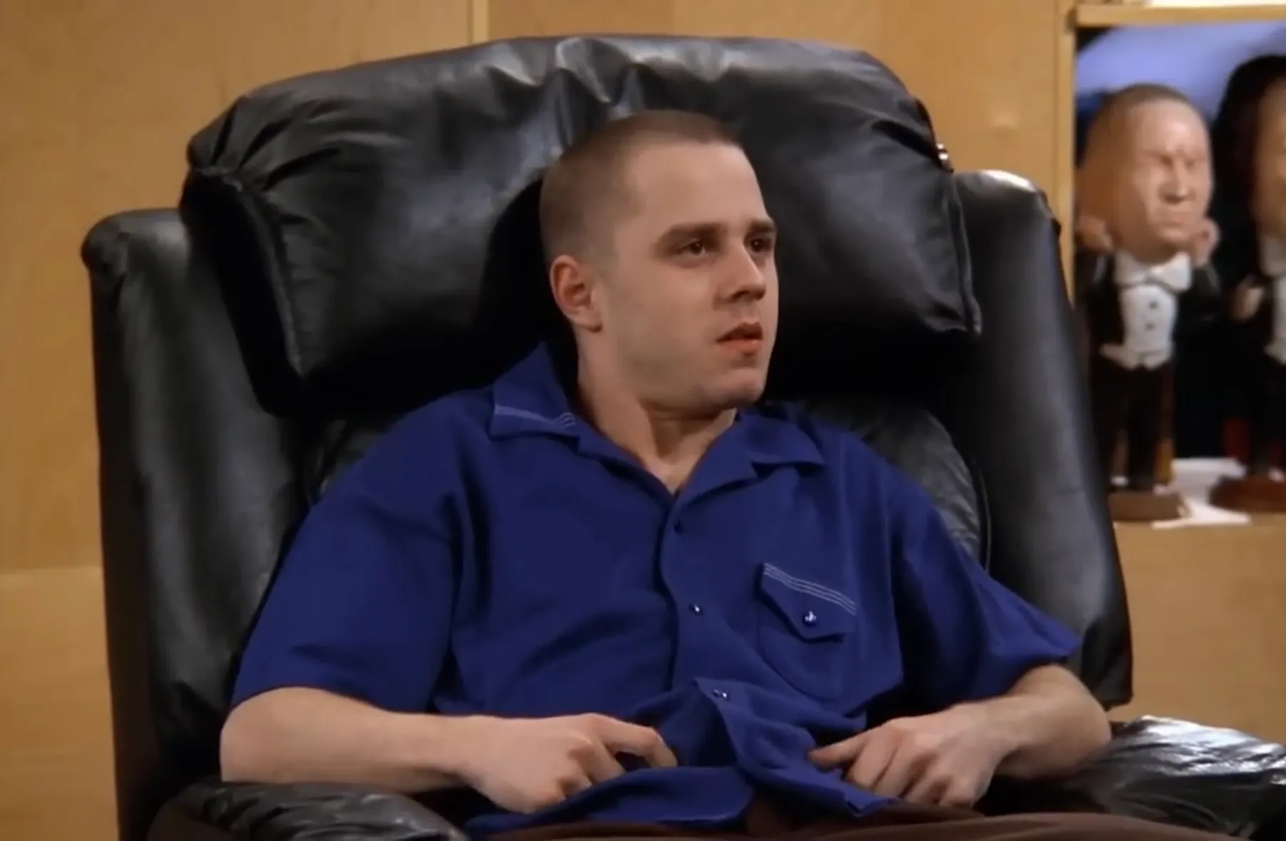 Frank Buffay Jr. from Friends sitting in a black leather chair wearing a blue shirt in an indoor setting.